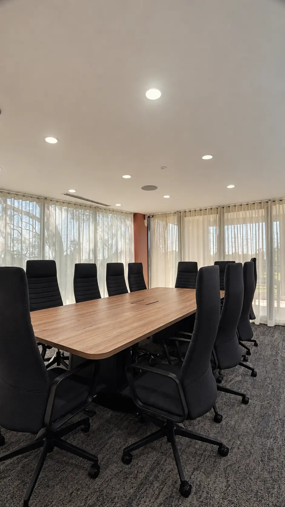 Board room with long timber table, black chairs and sheer curtains